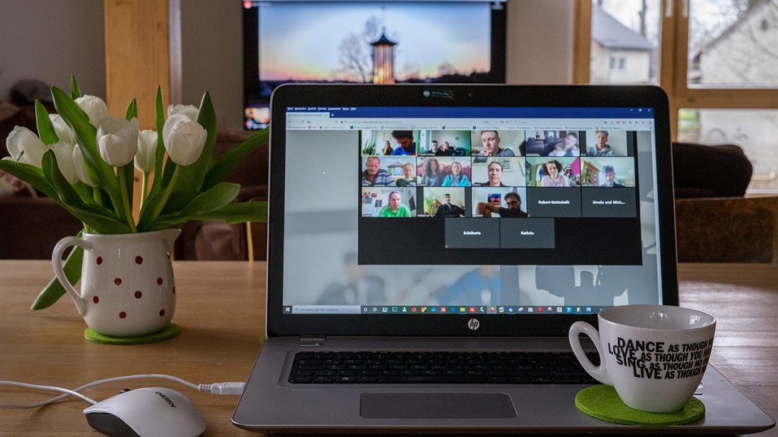 image shows a laptop screen with an active Zoom meeting, a vase of white flowers to the left and a coffee mug and mouse in the foreground.